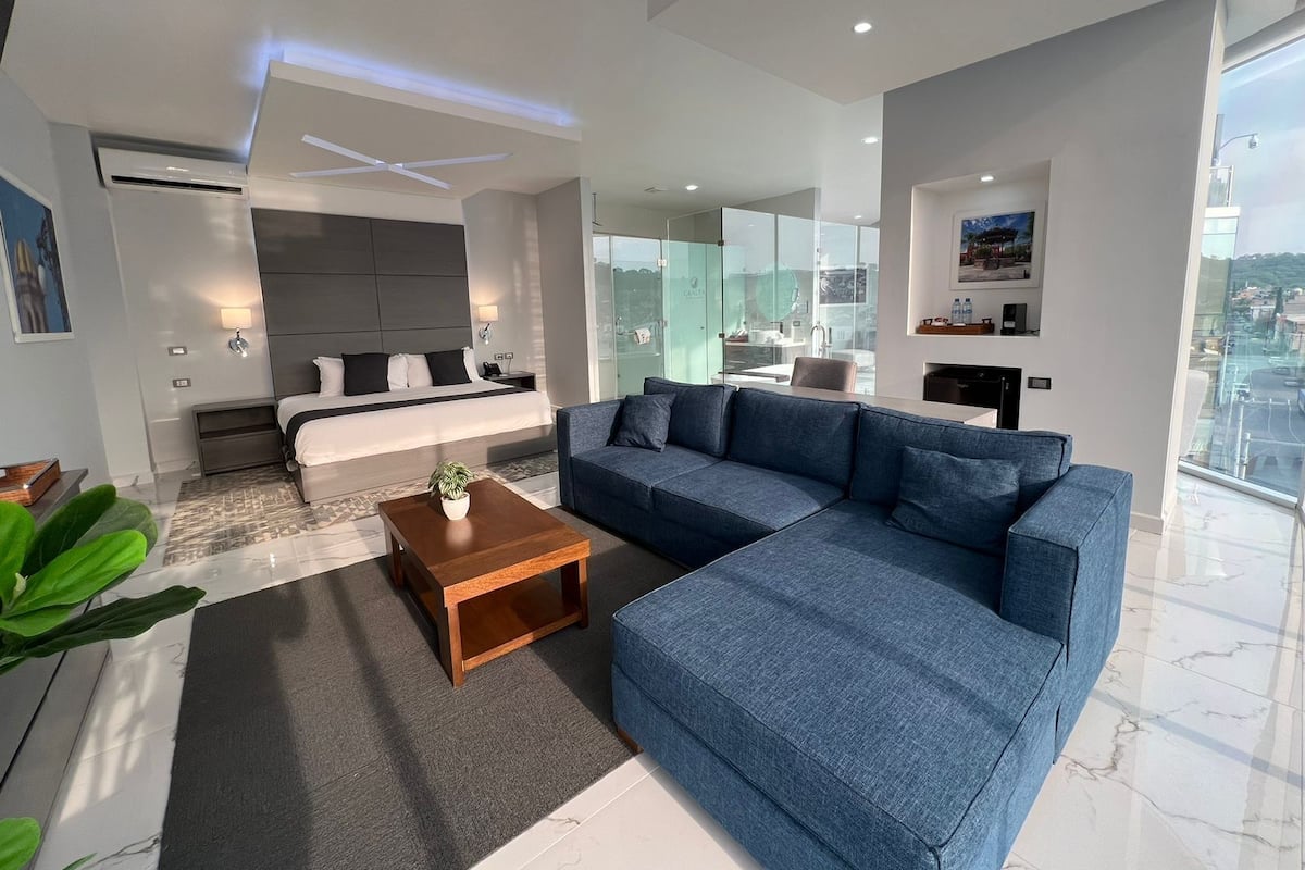 A modern hotel room with a large bed, a sofa, a coffee table, and a view of the city through large windows.