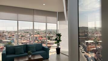 A spacious living room with a large window offering a city view, a teal sofa, a coffee table, and a potted plant.