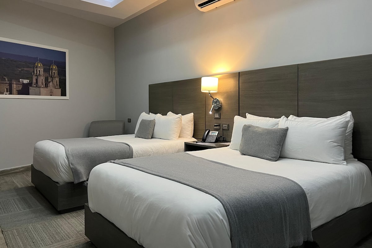 A hotel room with two beds, a nightstand with a lamp, an air conditioner, and a painting on the wall.