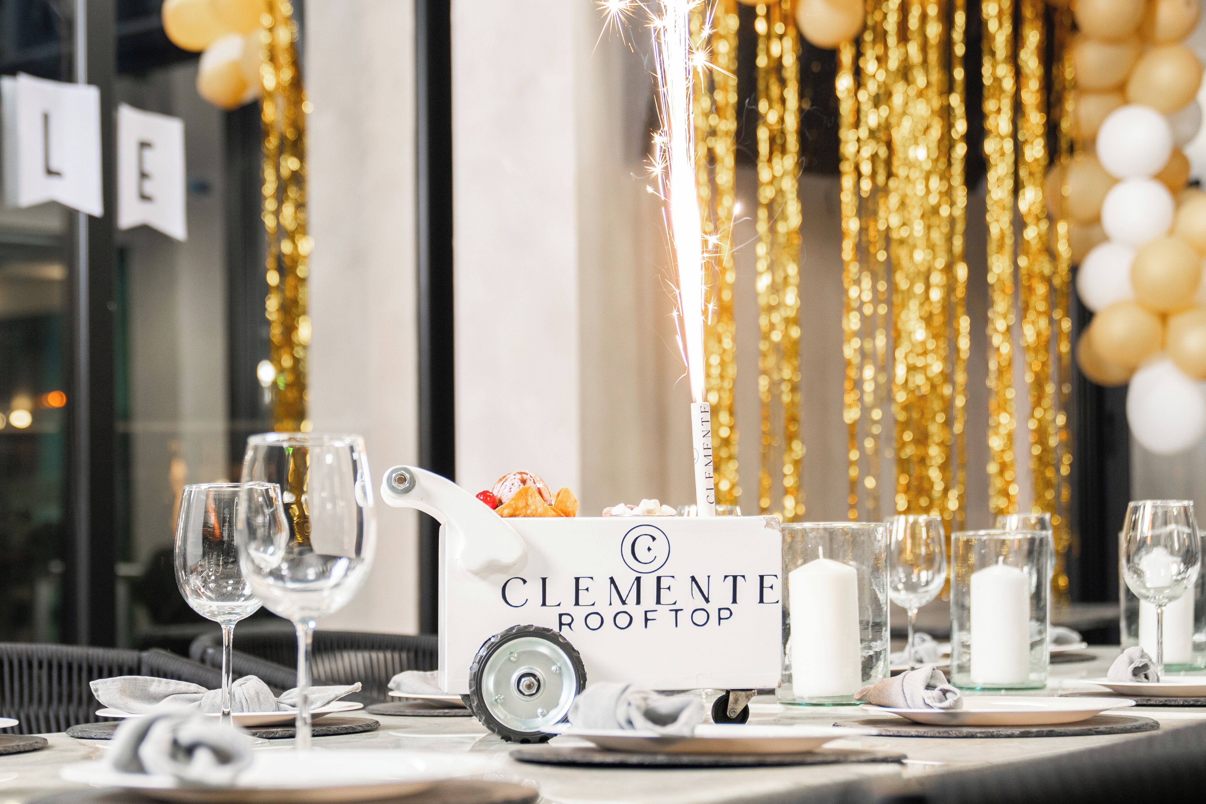 Une table dressée avec des verres à vin, un chariot portant l’inscription « Clemente Rooftop », et une bougie blanche.