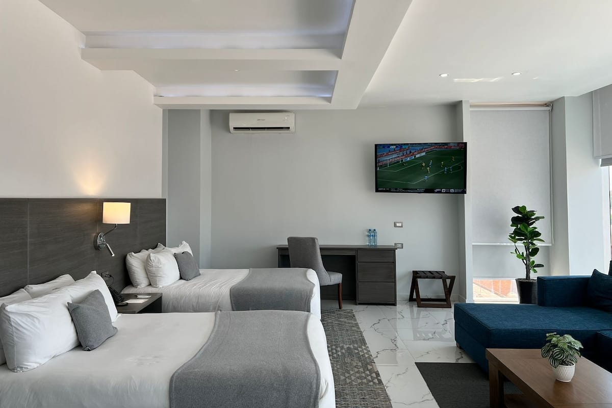 A modern hotel room with two beds, a desk, a TV, and a seating area.