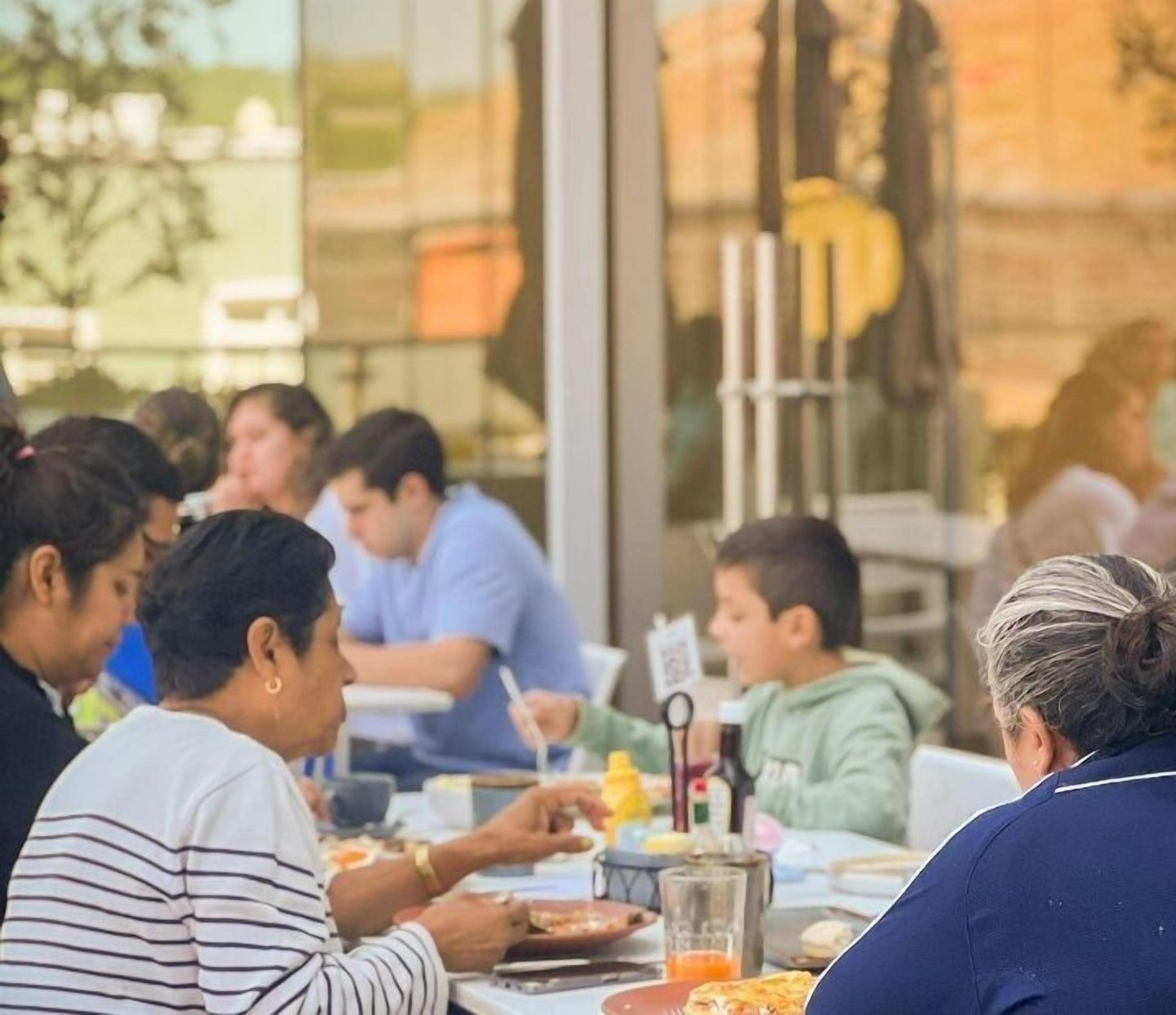 A group of people dining at an outdoor table with a glass partition, casual seating, and a view of the outdoors.