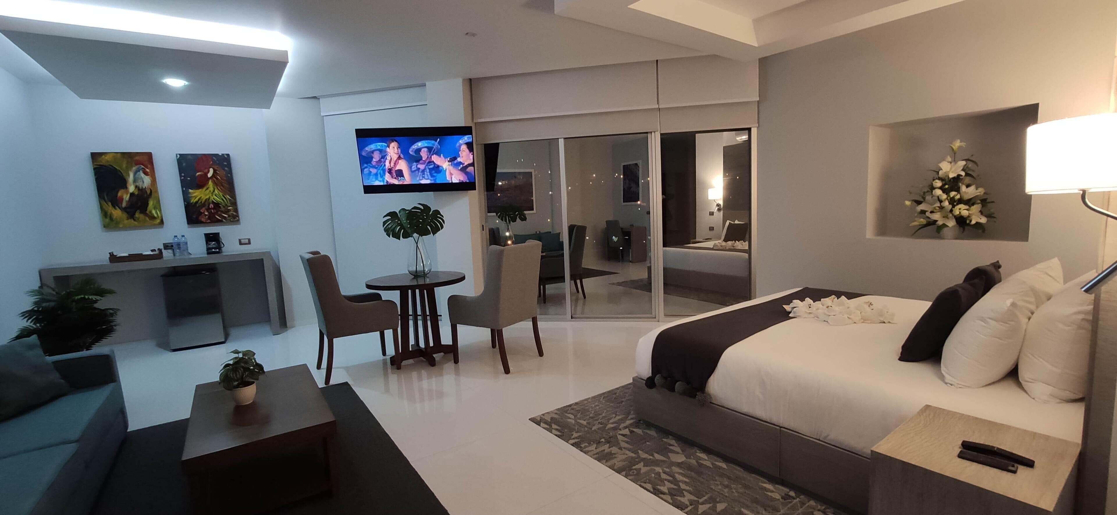 Habitación de hotel moderna con una cama grande, televisor, zona de estar con mesa y sillas, y balcón con vistas.