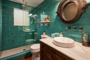 Un baño con mosaicos azules turquesa, inodoro blanco, ducha con mampara de vidrio, espejo redondo y lavamanos de mármol con artículos de aseo.