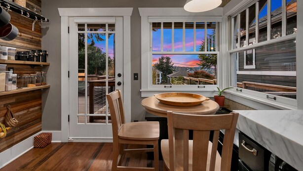 Una cocina con estantes de madera, una mesa de comedor con sillas y vistas a un atardecer a través de una ventana.