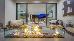 A modern living room with a glass coffee table featuring a fire feature, a sofa, and a dining area in the background. - Troy Dr - Sleek & Modern Hollywood Hills Home With Stunning Views (Los Angeles)