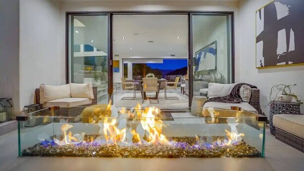 A modern living room with a glass coffee table featuring a fire feature, a sofa, and a dining area in the background. - Troy Dr - Sleek & Modern Hollywood Hills Home With Stunning Views (Los Angeles)
