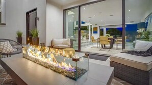 A modern outdoor seating area with a glass-top fire feature, wicker furniture, and a view of the interior through large glass doors. - Troy Dr - Sleek & Modern Hollywood Hills Home With Stunning Views (Los Angeles)