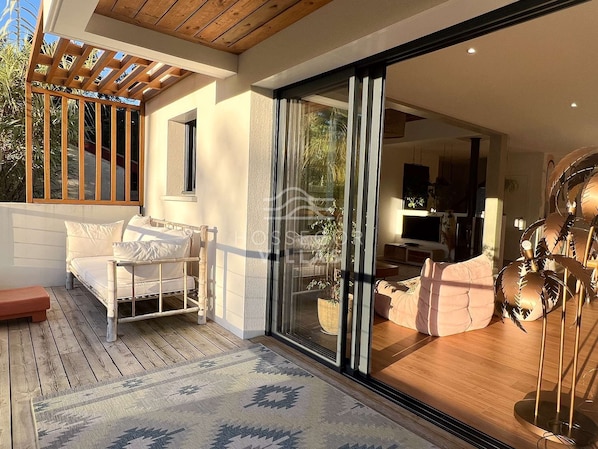 A modern interior with a wooden deck, a sofa, a dining area, and a sliding glass door leading to a garden.