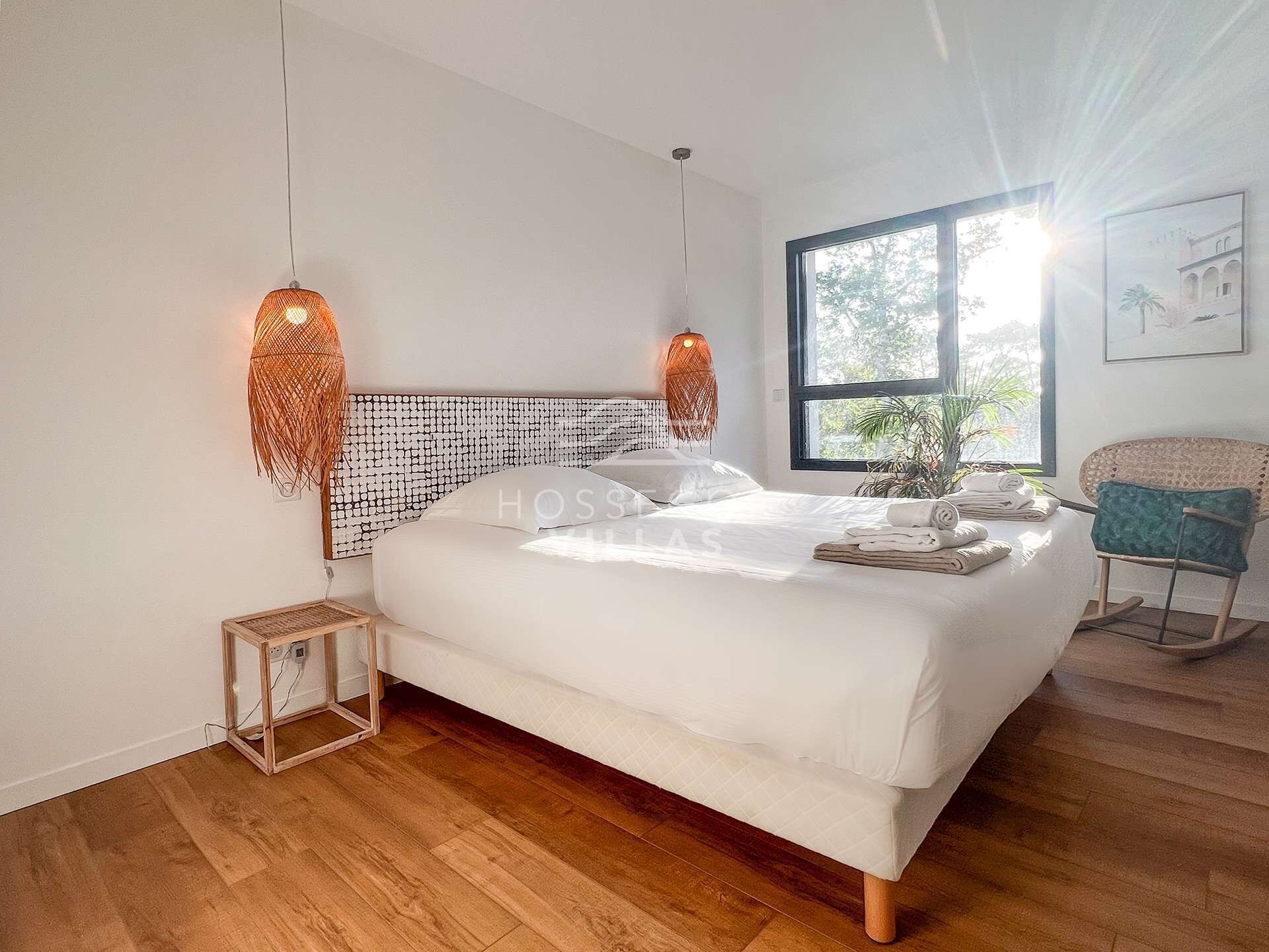 Ein modernes Schlafzimmer mit einem großen Bett, zwei hängenden Pendelleuchten, einem kleinen Nachttisch, einem Sessel und einem Fenster mit Blick auf Grünflächen.