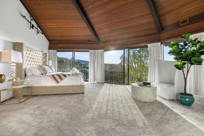 A bedroom with a large bed, a bedside table, a nightstand, a potted plant, and a view of the outdoors.