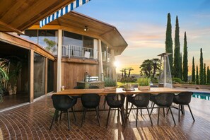 A modern outdoor dining area with a wooden table and chairs, a pool, and a view of tall trees and a sunset.