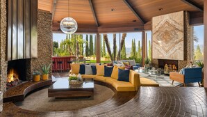 A modern living room with a curved sectional sofa, a fireplace, and a large window offering a view of trees and a distant landscape. - Via Vallarta - An Architecturally Significant California Modern Home (Encino)