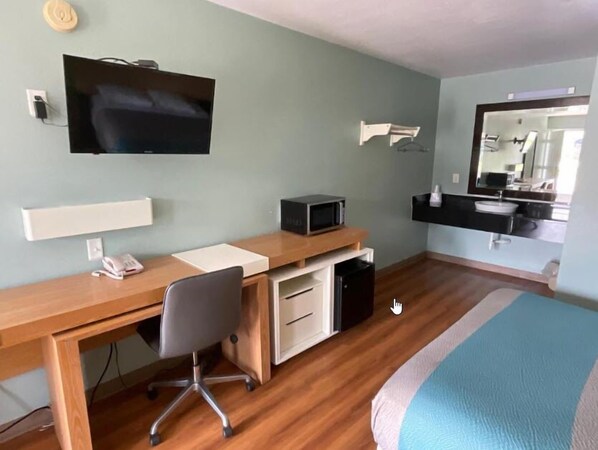A hotel room with a wooden desk, a television mounted on the wall, a microwave, a small refrigerator, a bedside table, and a bed with a blue and white striped bedspread.