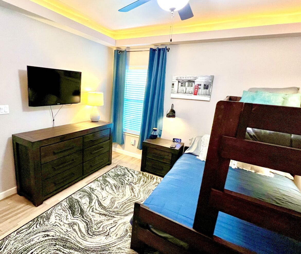 Un dormitorio con cama, cómoda, televisor, mesita de noche y ventilador de techo.