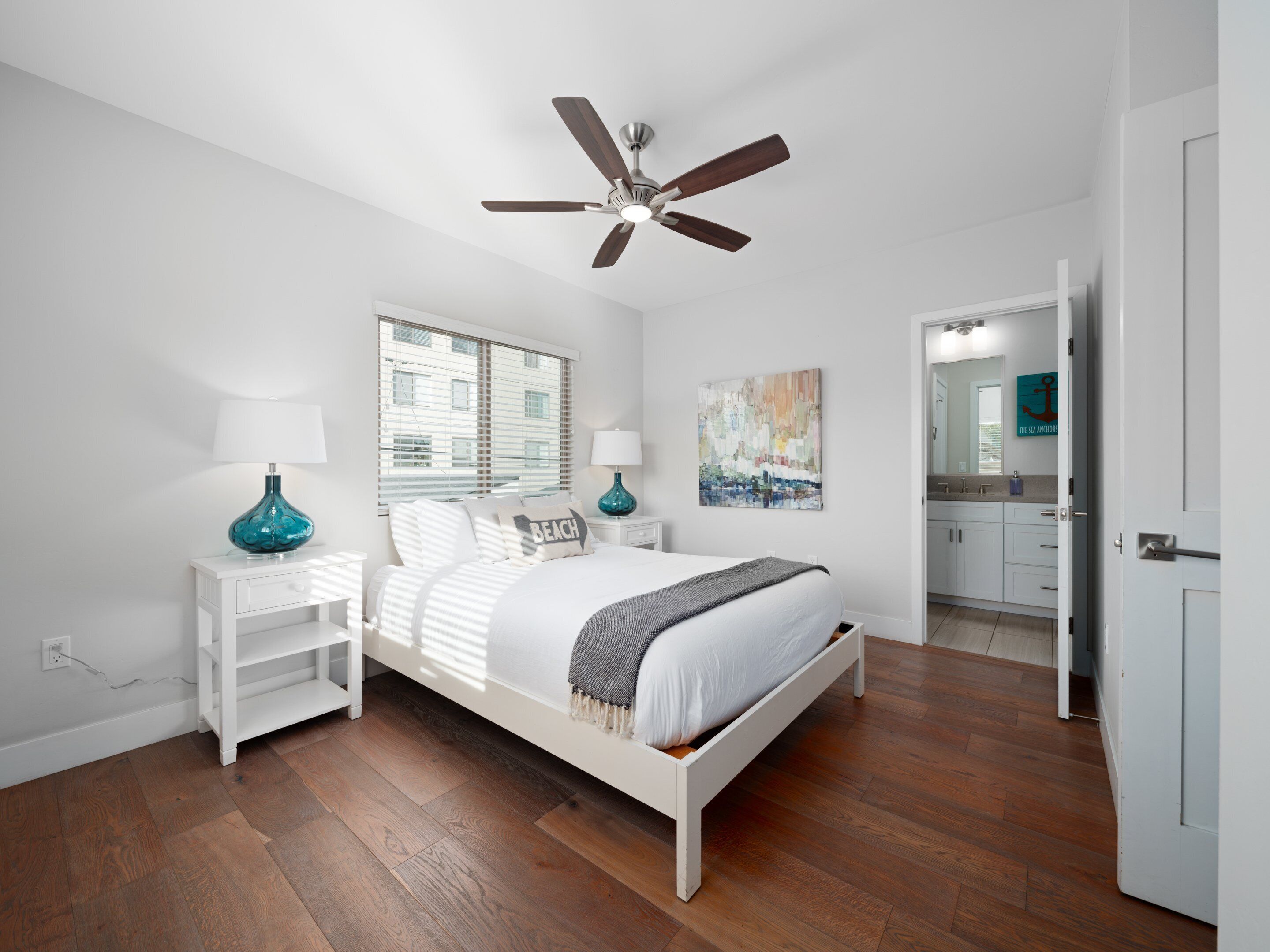 A bedroom with a bed, nightstands, a ceiling fan, a window with blinds, a painting, and a bathroom visible through an open door.