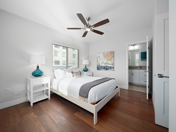 A bedroom with a bed, nightstands, a ceiling fan, a window with blinds, a painting, and a bathroom visible through an open door.