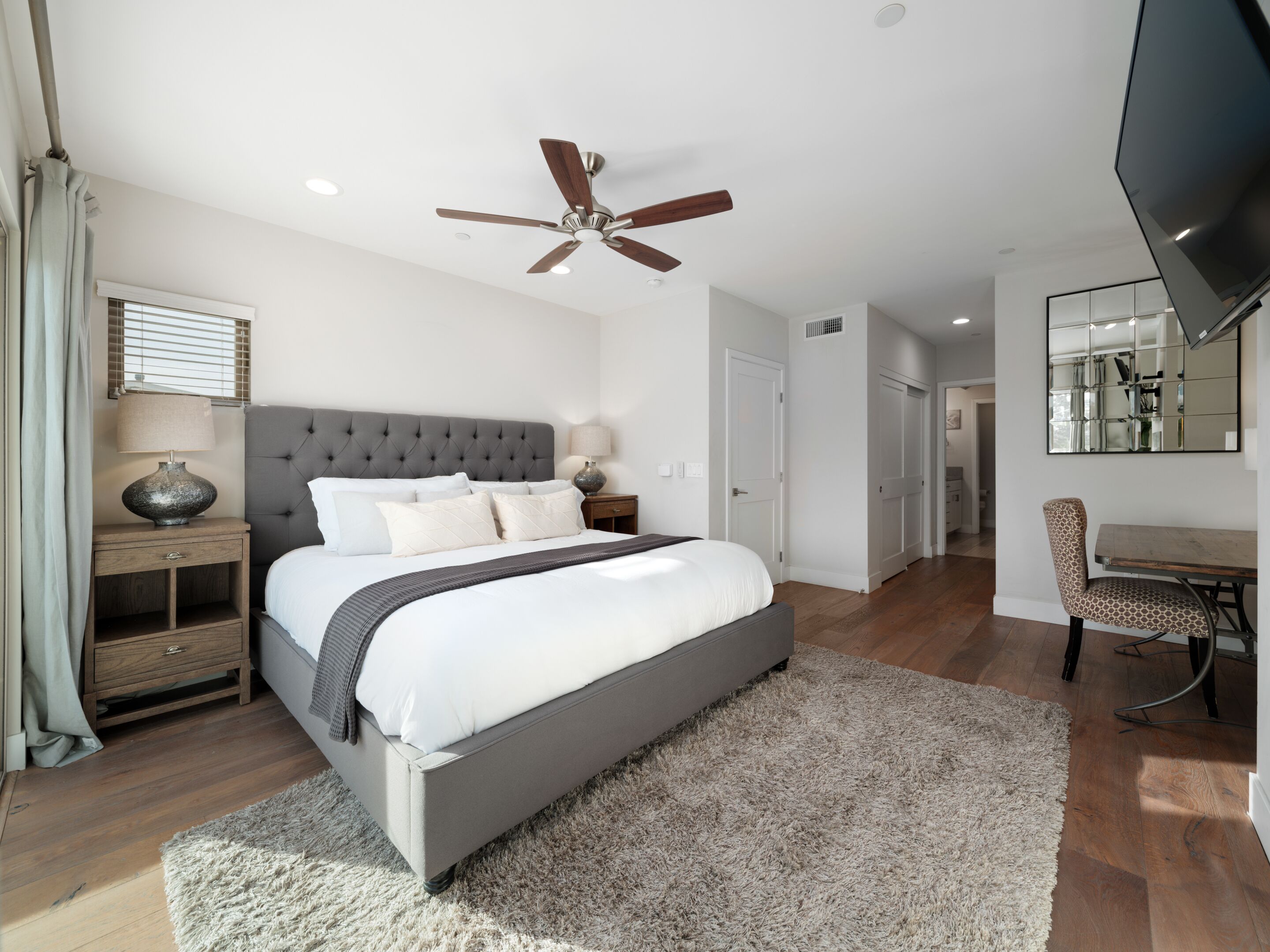 Une chambre moderne avec un grand lit, un ventilateur de plafond, une table de chevet avec une lampe et un téléviseur fixé au mur.