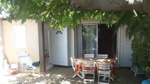 En terrasse med et bord og stole, en blå liggestol og en dør, der fører til et overdækket område.
