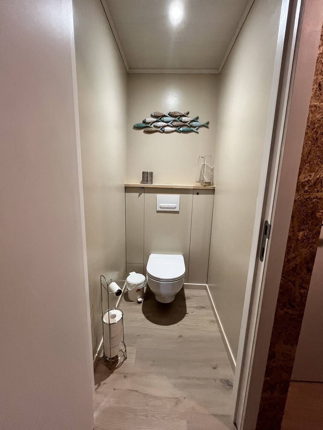 Une petite salle de bain comprenant des toilettes, une étagère avec des objets décoratifs et un luminaire fixé au mur.