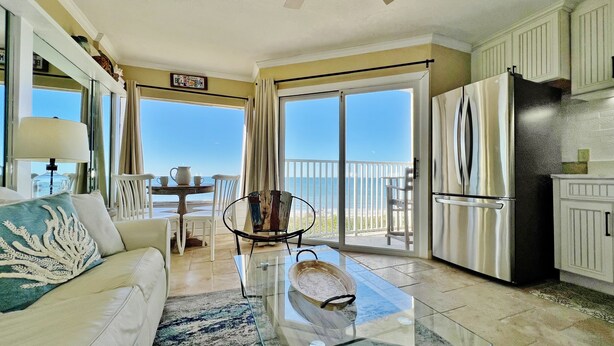 A spacious living area with a balcony, a kitchen with a stainless steel refrigerator, and a dining table with chairs. - Cozy Beachfront Retreat right on the Sand, Sparkling Pool, & Charming Coastal Flair! (Belleair Beach)