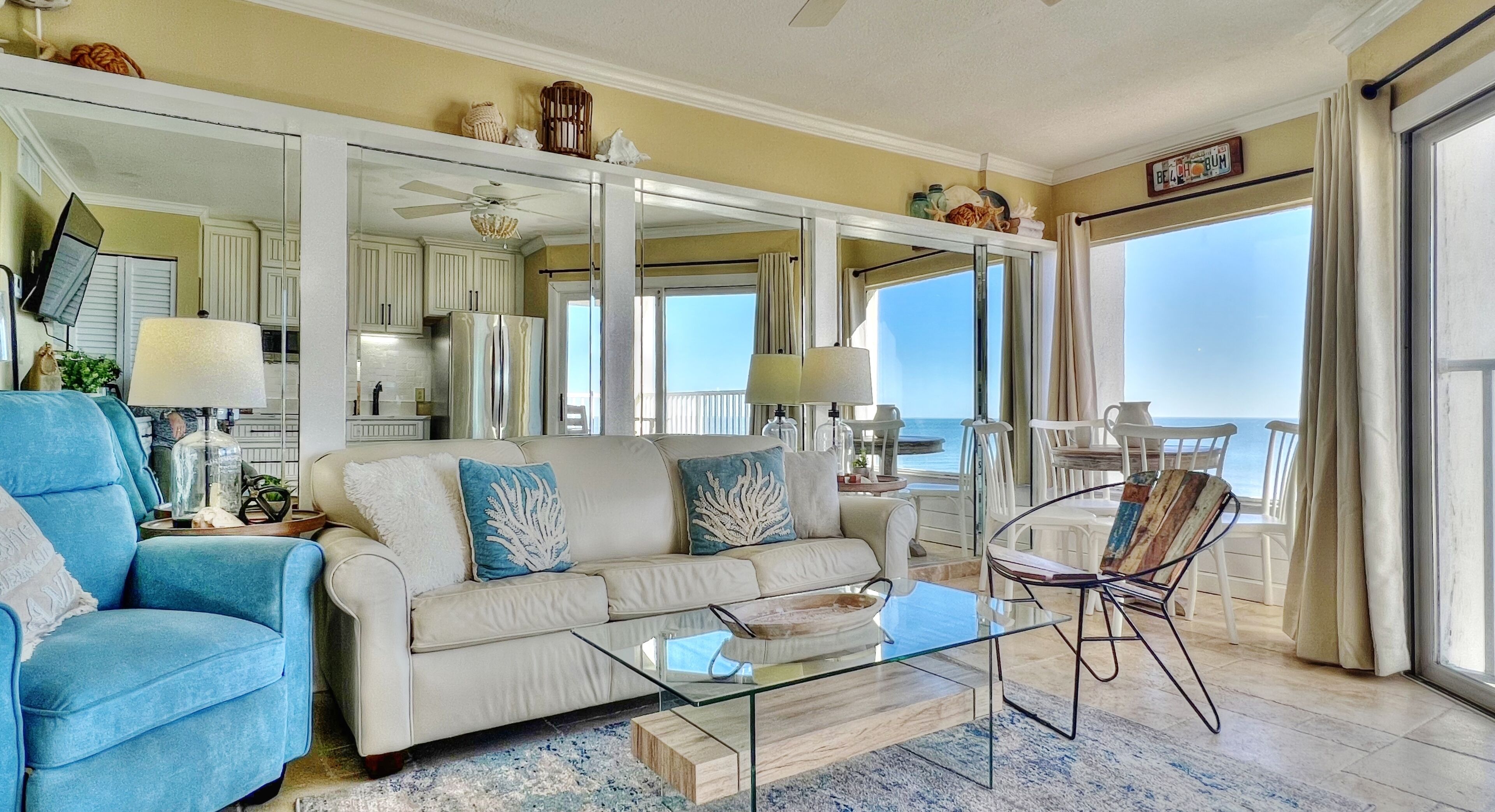 Un ampio soggiorno con un divano beige, una poltrona blu e un tavolino in vetro. Un grande specchio riflette la stanza e un balcone con vista sull'oceano.