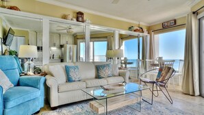 A spacious living room with a beige sofa, a blue armchair, and a glass coffee table. A large mirror reflects the room and a balcony with a view of the ocean. - Cozy Beachfront Retreat right on the Sand, Sparkling Pool, & Charming Coastal Flair! (Belleair Beach)