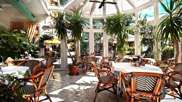 A restaurant with wicker chairs and tables, surrounded by potted plants and a glass ceiling.