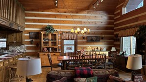 Ein gemütliches Blockhaus-Interieur mit einer Essecke aus Holz, einer Küche mit einem großen Holzkasten und einem Wohnzimmer mit einer Couch und Zierkissen.