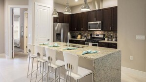 Une cuisine moderne avec des meubles de couleur sombre, des appareils en acier inoxydable et un comptoir en granit. On y trouve quatre tabourets de bar blancs et des luminaires suspendus au-dessus de l’îlot central.