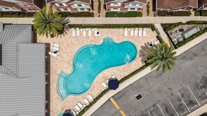 Vista aerea di una piscina con lettini e ombrelloni, circondata da un parcheggio e edifici residenziali.