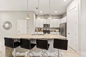 A modern kitchen with a granite island, black bar stools, white cabinets, stainless steel appliances, and pendant lighting. - 4BR Summerville Resort Villa in Kissimmee | Near Disney, Pool, Safety (Kissimmee)