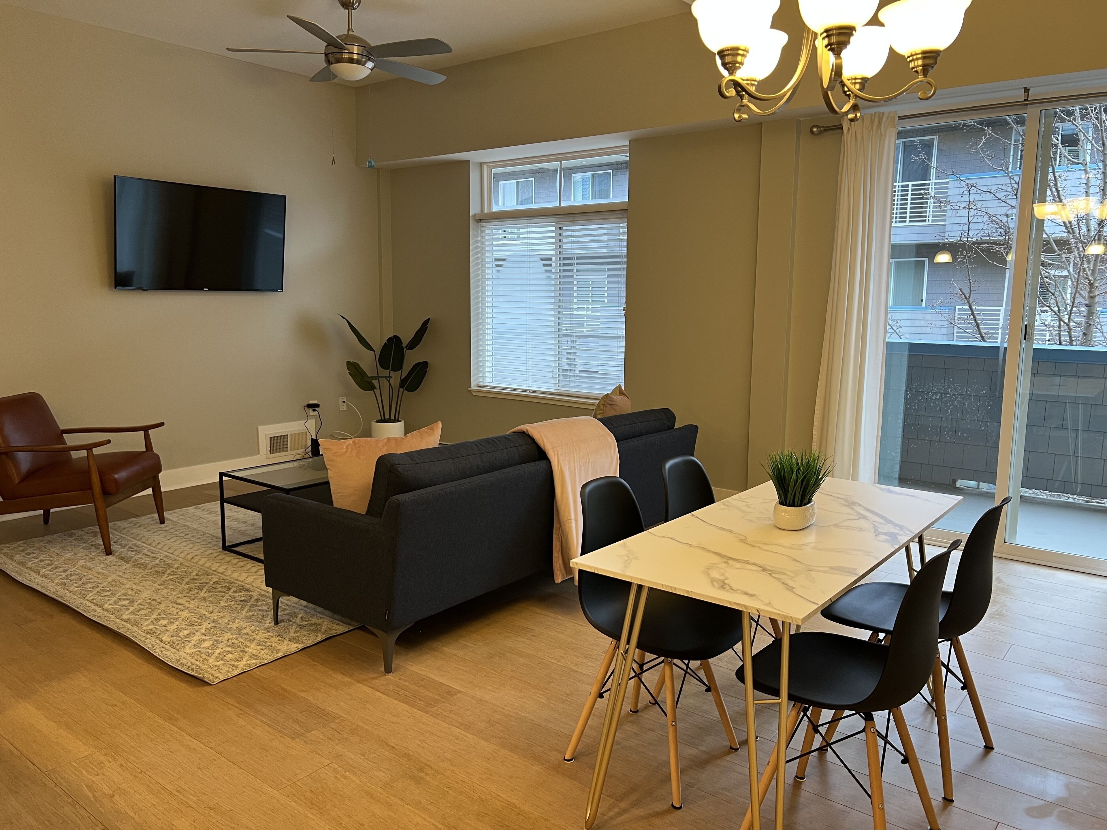 A modern living room with a sofa, a dining table with chairs, a TV mounted on the wall, and a ceiling fan.