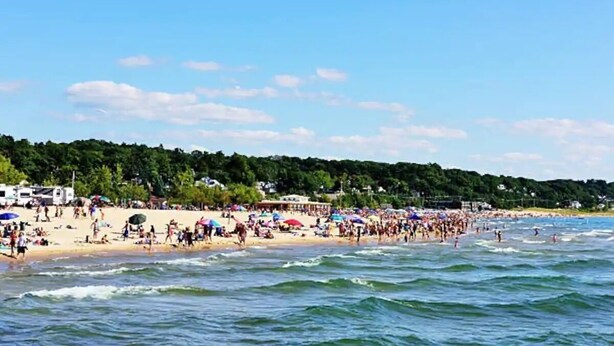 Ein belebter Strand mit Menschen und Sonnenschirmen, ein klarer Himmel und ein bewaldeter Hügel im Hintergrund.