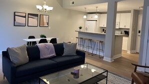 Ein modernes Wohnzimmer mit einem dunklen Sofa, einem Glastisch und einem Essbereich mit weißem Tisch und Stühlen.