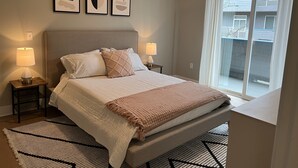 Ein Schlafzimmer mit einem Bett, zwei Nachttischen mit Lampen, einem Fenster mit Vorhängen und drei gerahmten Kunstwerken an der Wand.