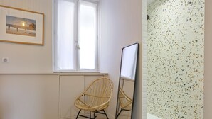 Une pièce dotée d’une grande fenêtre, d’un tableau encadré au mur, d’un fauteuil en osier, d’une petite table et d’un mur à texture.