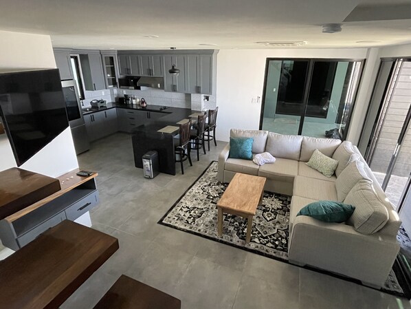 A modern living room with a sofa, coffee table, and a dining area with chairs. A kitchen with grey cabinets and a black countertop is visible in the background.