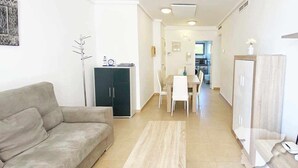 Een woonkamer met een bank, een eettafel met stoelen, een keuken op de achtergrond en een kapstok.