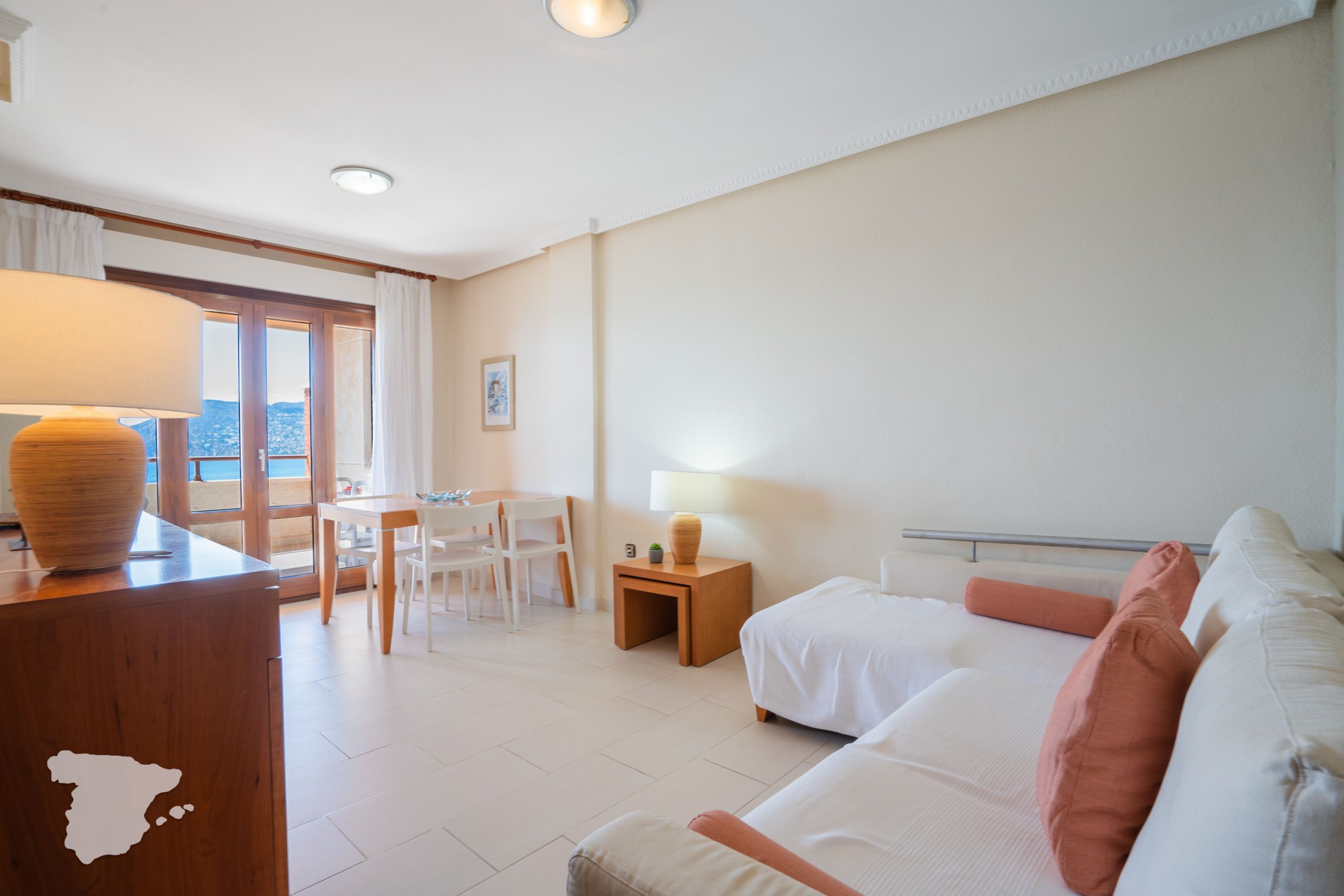 Habitación de hotel con escritorio de madera, sofá blanco, una mesita con lámpara y vista al mar a través de un ventanal grande.