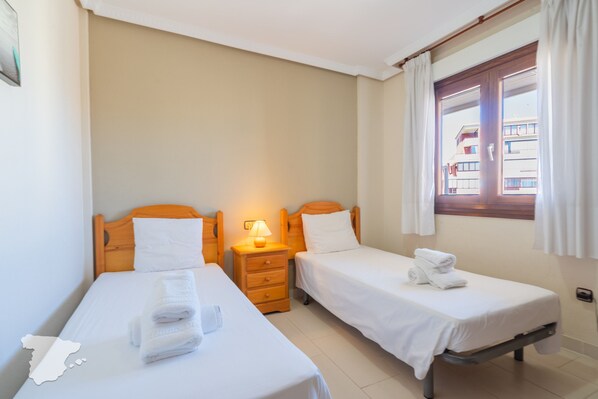 A hotel room with two single beds, a nightstand with a lamp, and a window with curtains.