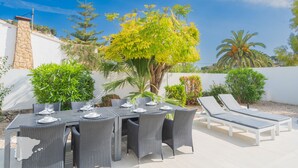 Un espacio de comedor al aire libre con una mesa dispuesta para seis personas, rodeado de sillas de mimbre y dos tumbonas.
