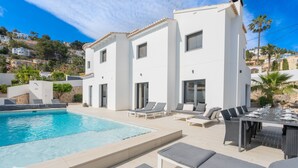 Una villa moderna con piscina, mobiliario exterior y vistas a la colina circundante.