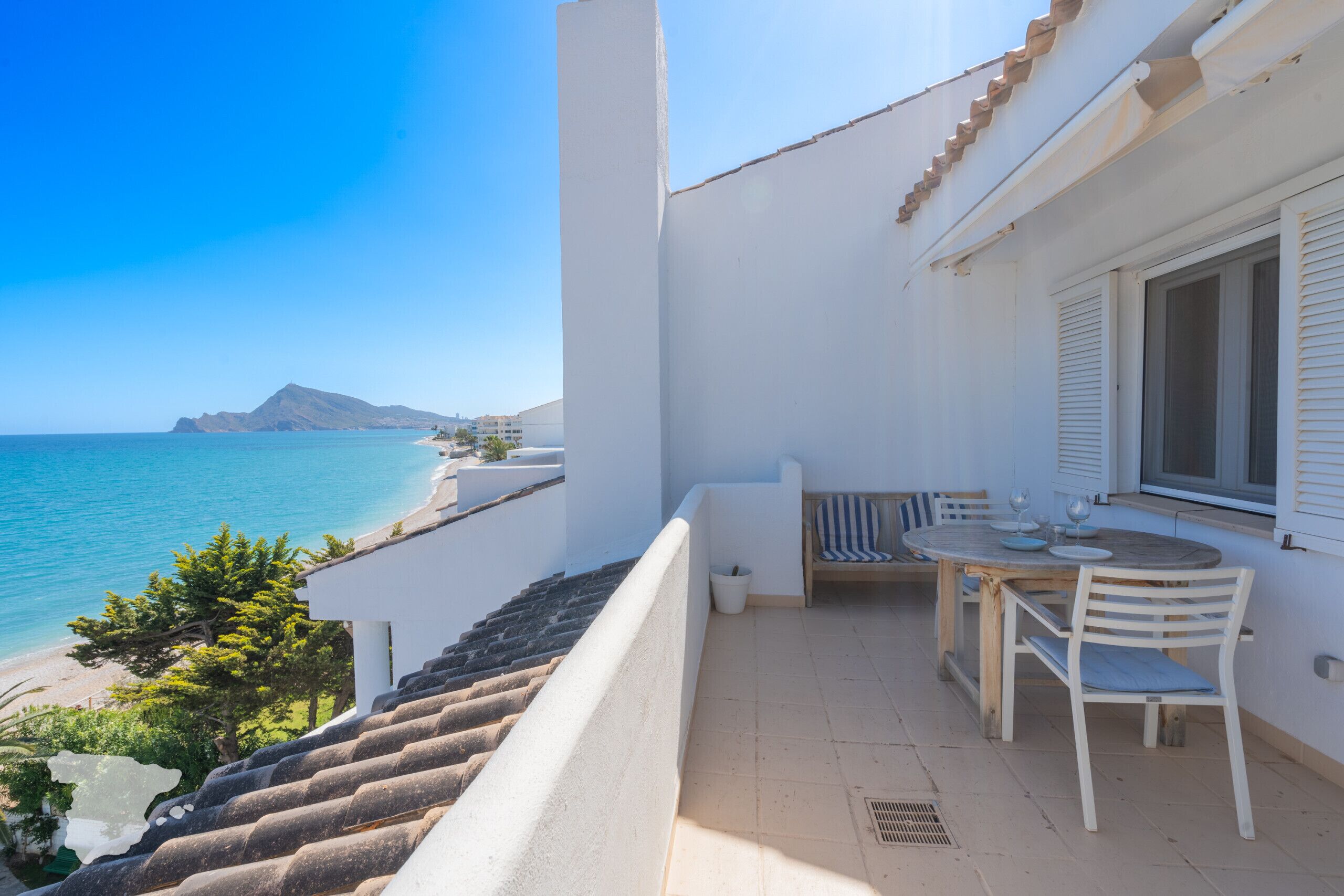 A balcony with a table and chairs, overlooking a beach and the sea.