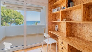 A room with a wooden bookshelf, a white chair, and a sliding glass door leading to a balcony with a view of the sea and trees. - CostaBlancaDreams - Duplex Cap Negret in Altea (Altea)