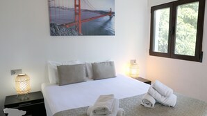 Un lit bien fait, avec des draps et des oreillers blancs, une fenêtre donnant sur des arbres et un tableau représentant le Golden Gate Bridge accroché au mur.
