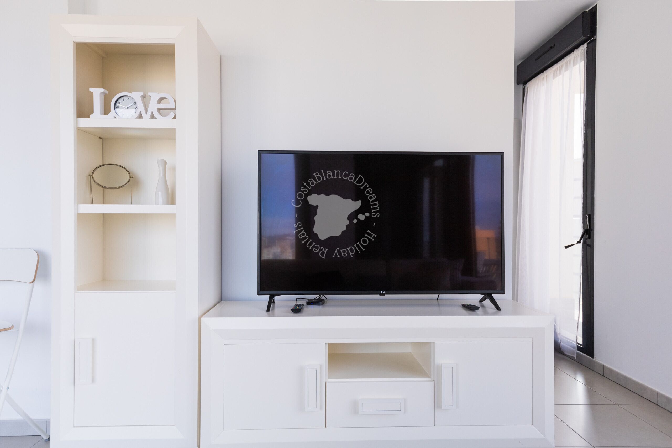 A modern living room with a flat-screen TV on a white entertainment unit, a white bookshelf with decorative items, and a large window with sheer curtains.