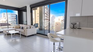 Ein modernes Wohnzimmer mit einer Couch und einem Couchtisch, großen Fenstern mit Blick auf die Stadt sowie einer Küchenzeile mit weißen Schränken und einer schwarzen Kücheninsel.