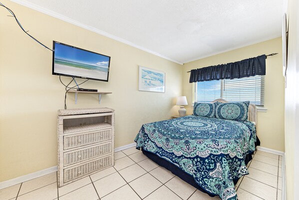 A bedroom with a bed, a television mounted on the wall, a framed picture, and a window with blinds.