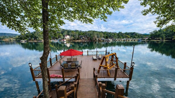 A wooden dock leading to a calm lake with a table and chairs, surrounded by trees and houses in the background.